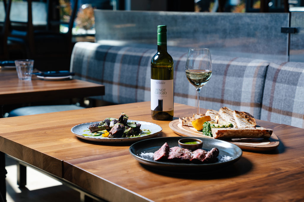 Wine bottle and glass on a wooden table with plates of steak, bread, and vegetables in a restaurant setting.