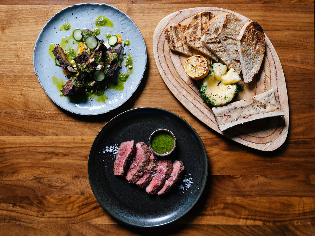 Sliced medium-rare steak on a black plate with green herb sauce and a small cup on a wooden table.