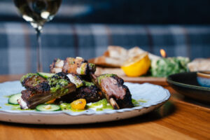 Plate of grilled ribs with herb sauce, cucumber slices, and yellow tomatoes, with a wine glass and bread in the background on a wooden table.
