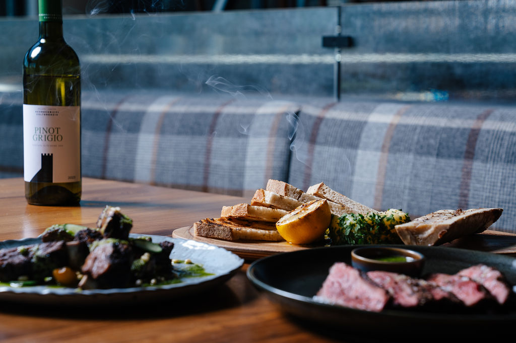 Wine bottle beside plates of bread, lemon, and herb-topped dishes on a wooden table at a dining setup under indoor lighting