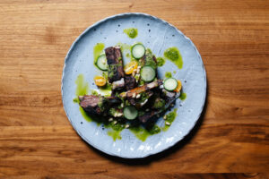 Plate of sliced grilled beef with green herb sauce, cucumber rounds, corn kernels, and sesame seeds on a blue plate