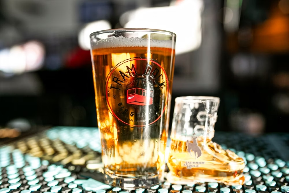 Tall amber beer in a pint glass with a circular tram logo on the front, set on a bar counter.