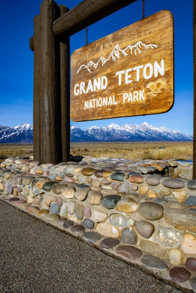 Wooden Grand Teton National Park sign hanging from a frame with snow-capped mountains in the background under a clear blue sky.