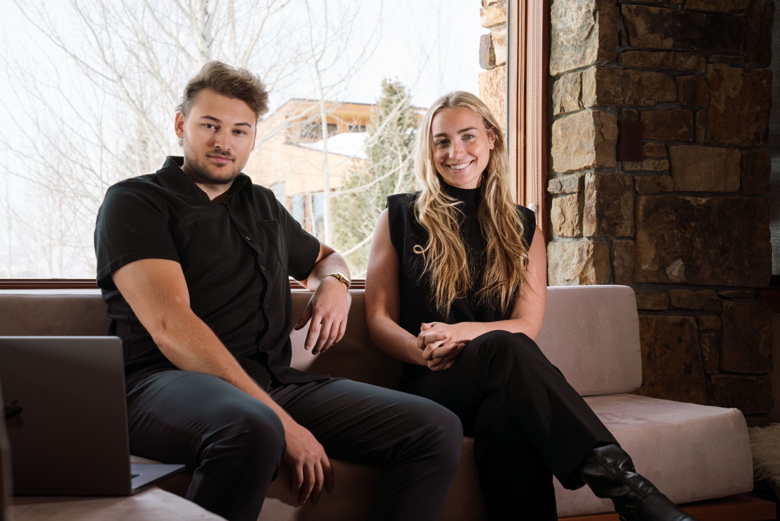 Man and woman sit on a beige couch by a stone wall, smiling at the camera with a laptop nearby outside the window.