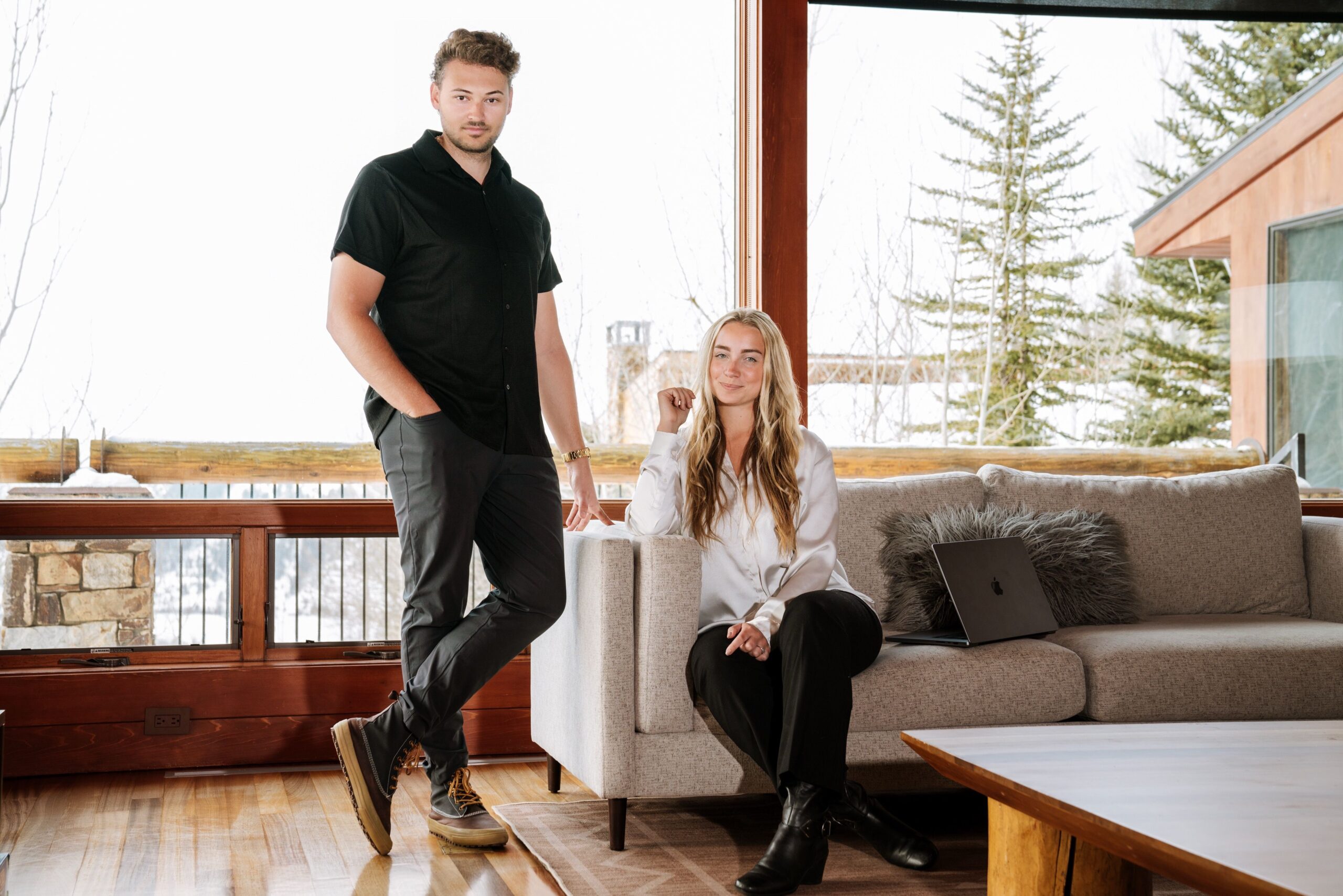 Man stands beside a light gray sofa as a woman sits nearby with a laptop, in a bright living room with wooden accents and large windows outside.