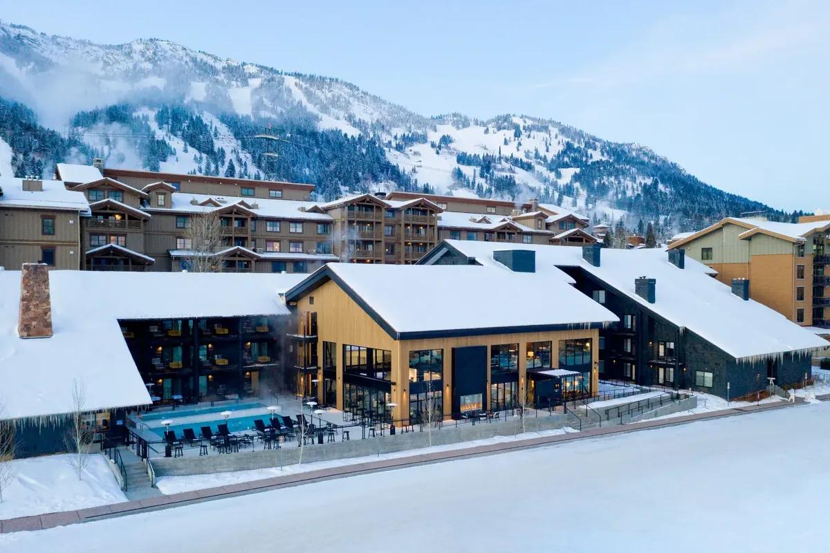 A snow-covered mountain lodge with a heated outdoor pool, steam rising, below forested ski slopes.