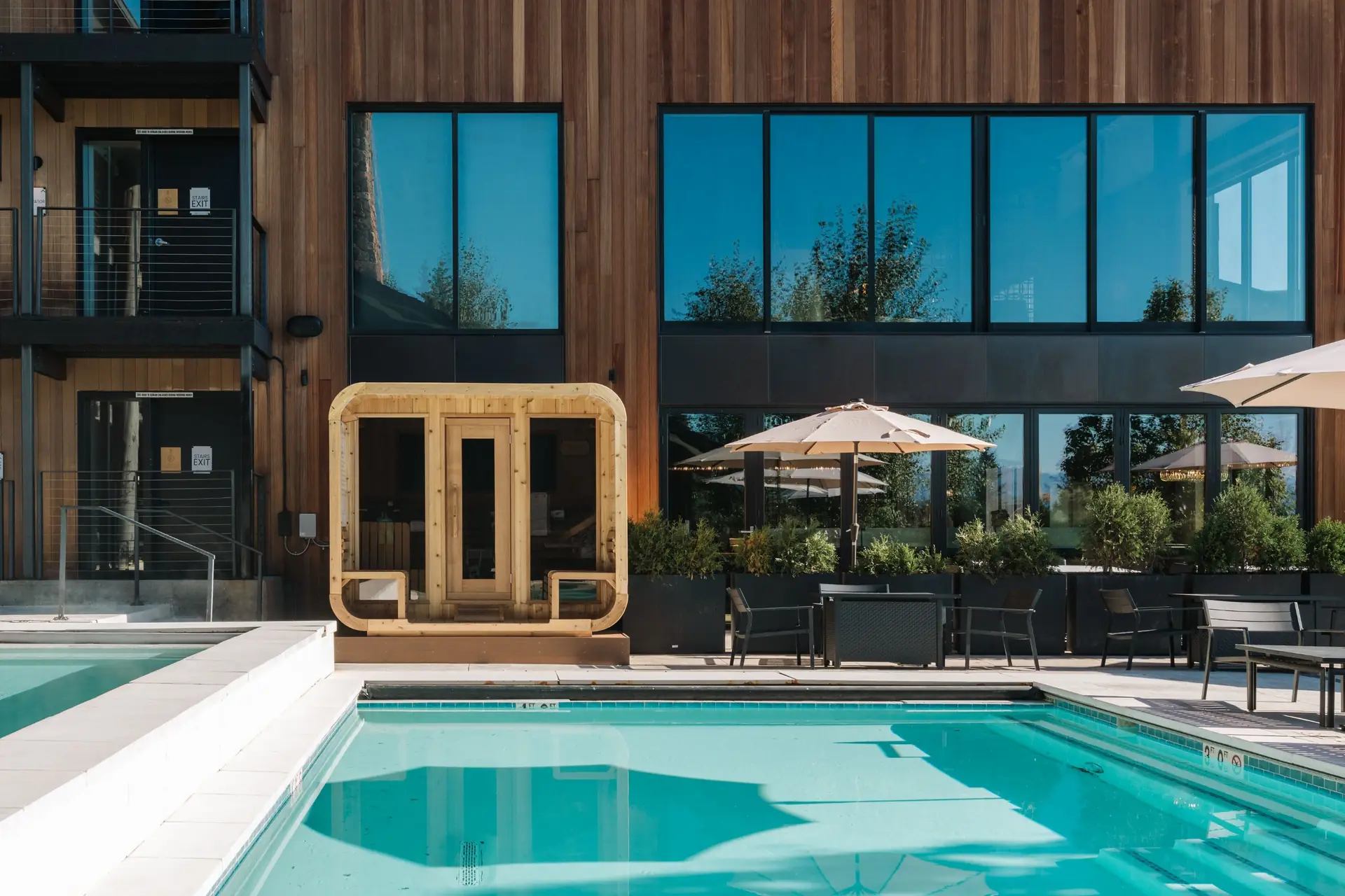 A wooden sauna beside an outdoor pool, in front of a wood-clad building with tall black windows and a patio with umbrellas.