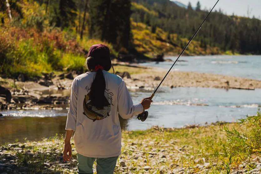A riverbank in autumn colour, with The Riverbend Project badge on a fishing shirt and a fly rod, and forested hills beyond.