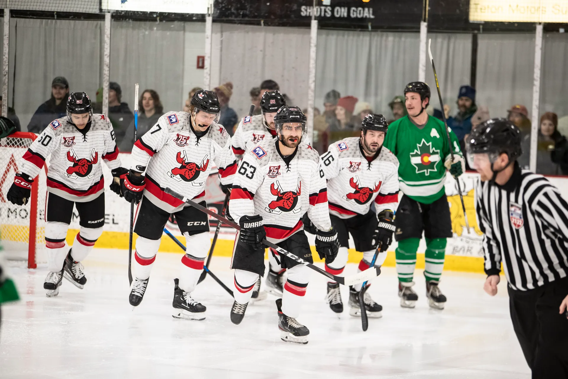 Hockey players in white and red jerseys skate across the ice, with a referee in the foreground and fans behind the glass.