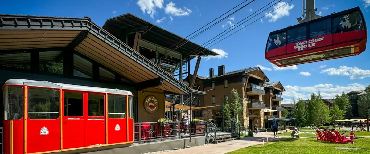 A red vintage tram cabin by the restaurant's deck on a summer day, the aerial tram overhead and red chairs on the lawn.