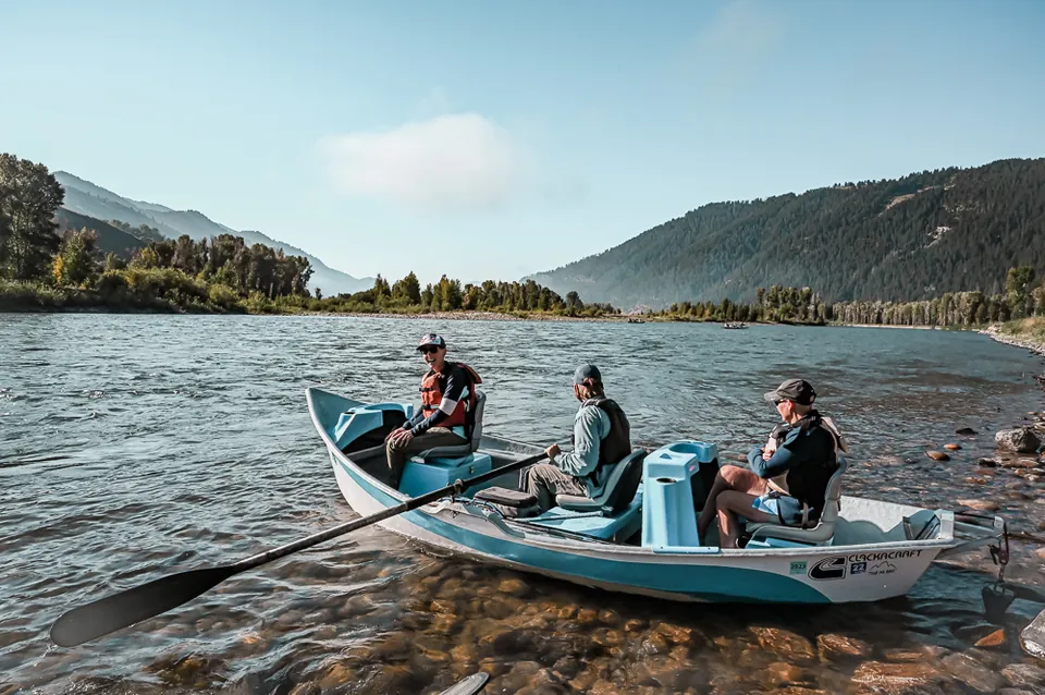 Three anglers in a drift boat on a wide river, with forested mountains rising behind.