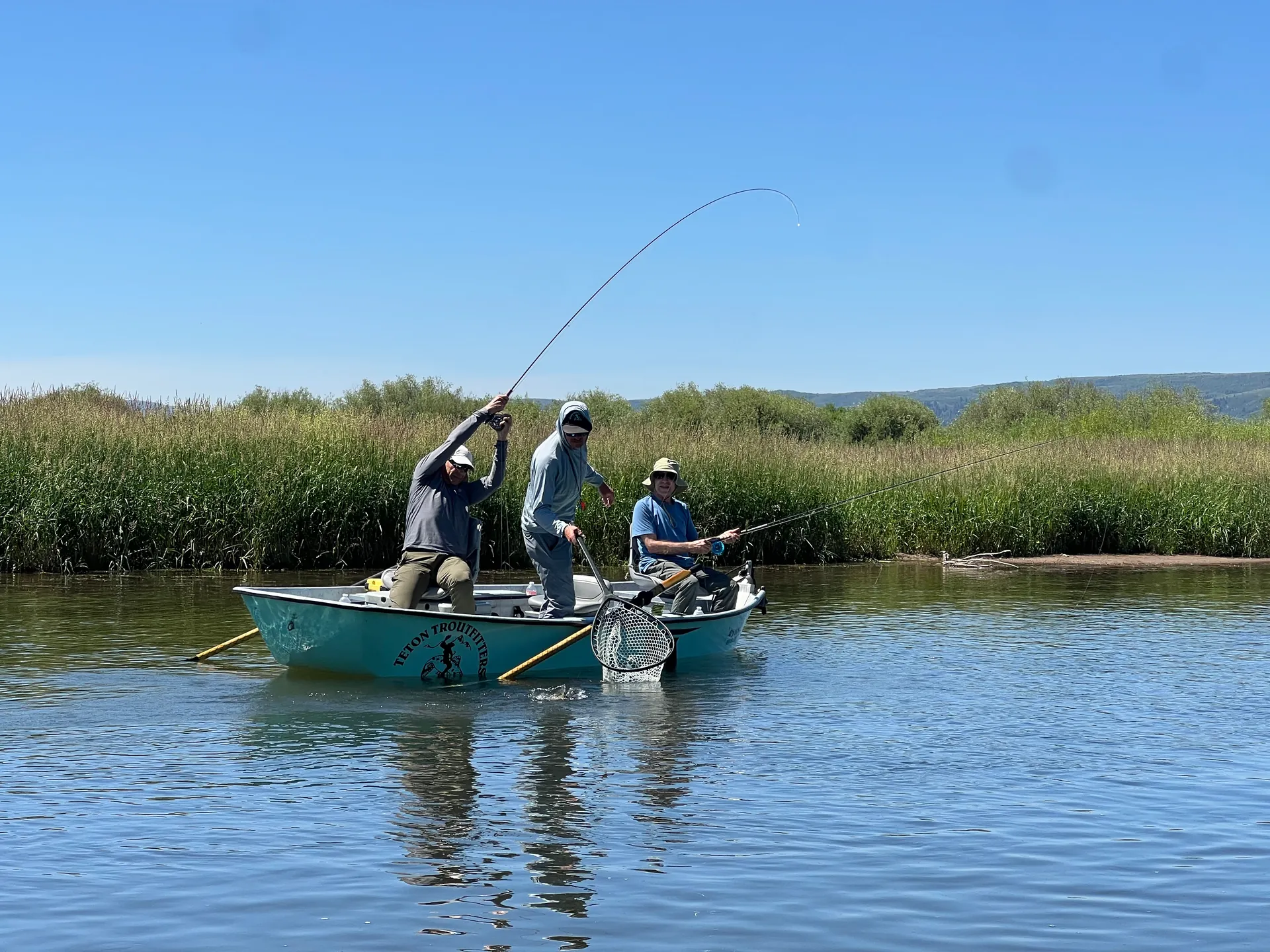 Three anglers fish from a drift boat on a reed-lined river, one rod bent into a fish.