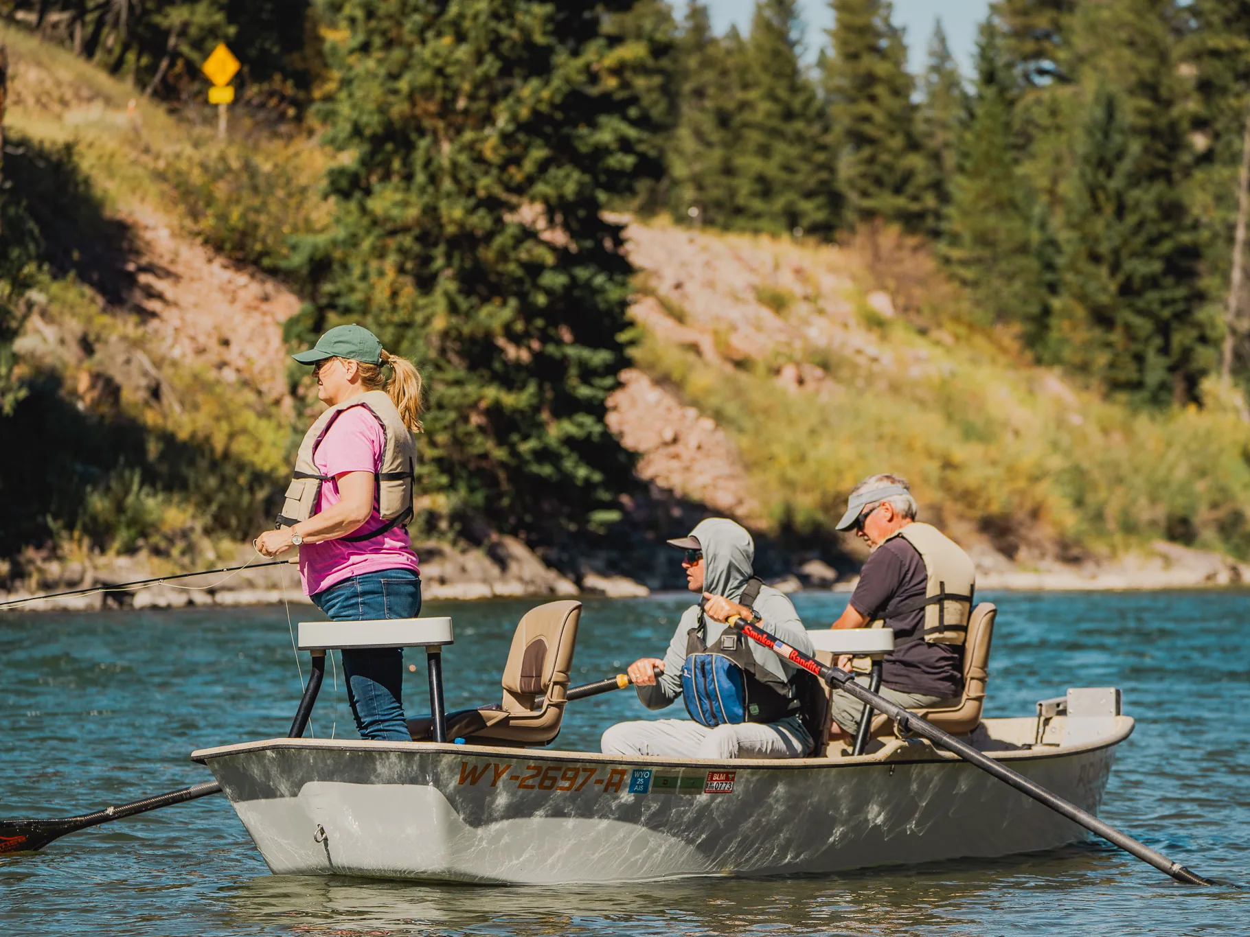 Three people in a drift boat on a river: one stands at the bow to cast, one rows, one sits at the stern, wooded hills behind.