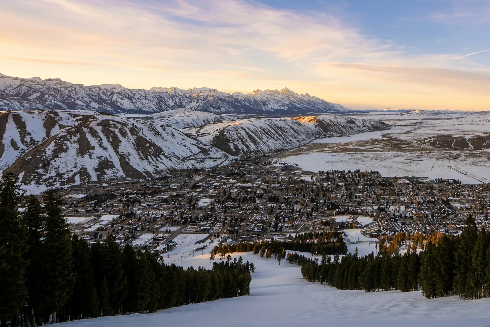 Jackson from above at golden hour: a snowy ski run leads down to the town, snow-covered hills and a mountain range behind.