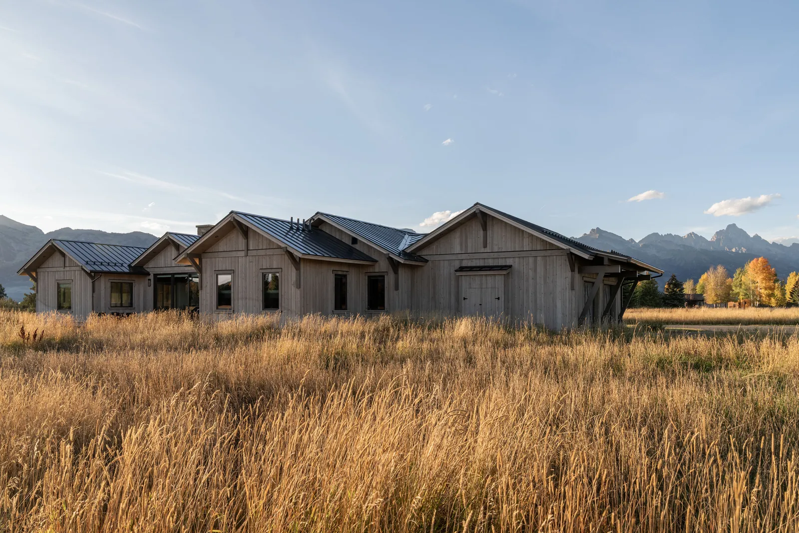 A single-story home clad in weathered gray wood, set in tall golden grass, with jagged peaks behind in evening light.