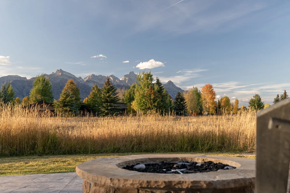 A stone fire pit on a patio, looking across a meadow of tall grass and autumn trees to a jagged mountain range.