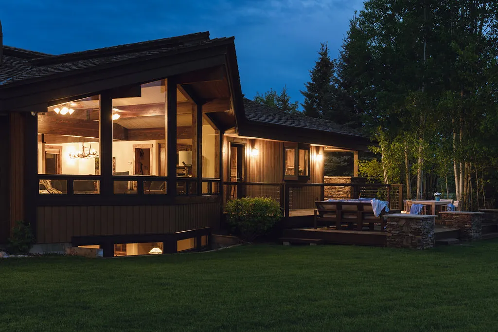 A wood-sided home at dusk, its tall corner windows lit from inside, beside a deck with stone pillars and a stand of aspens.