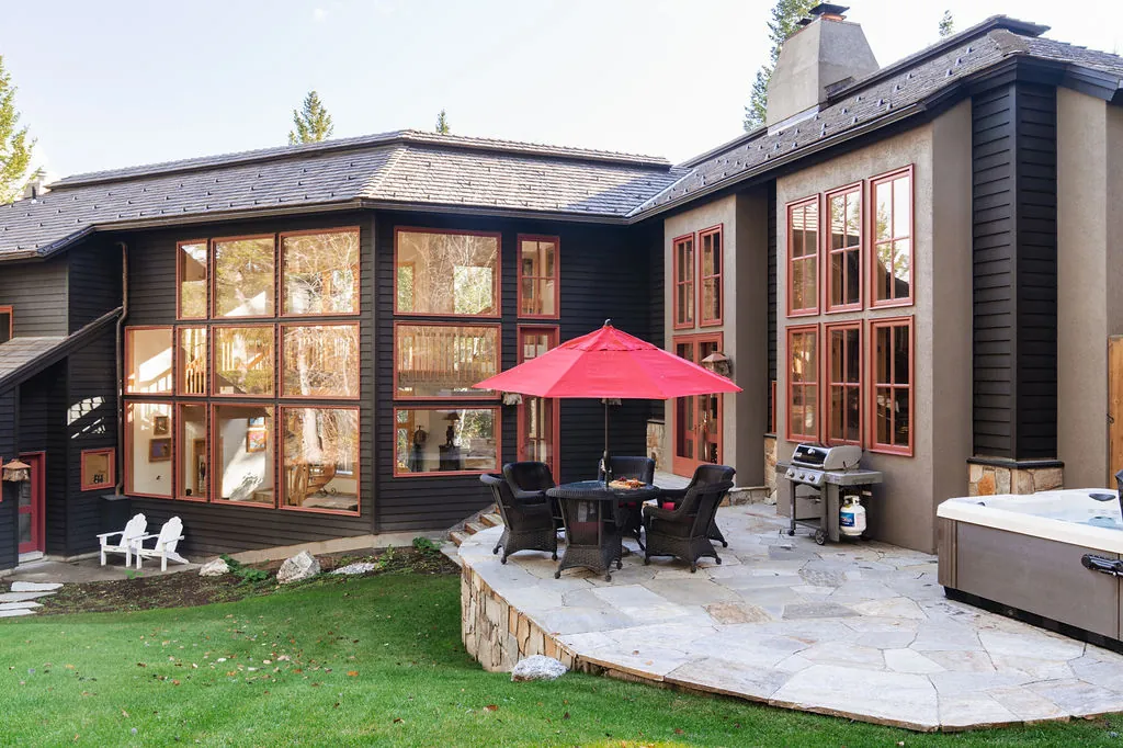 The back of a dark-sided home with tall red-framed windows, and a flagstone patio with a red umbrella, a grill and a hot tub.