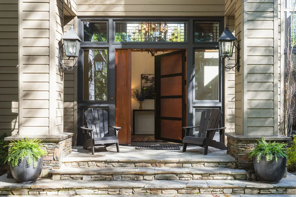 An open wooden front door between tall windows, with two Adirondack chairs, lanterns and potted ferns on stone steps.