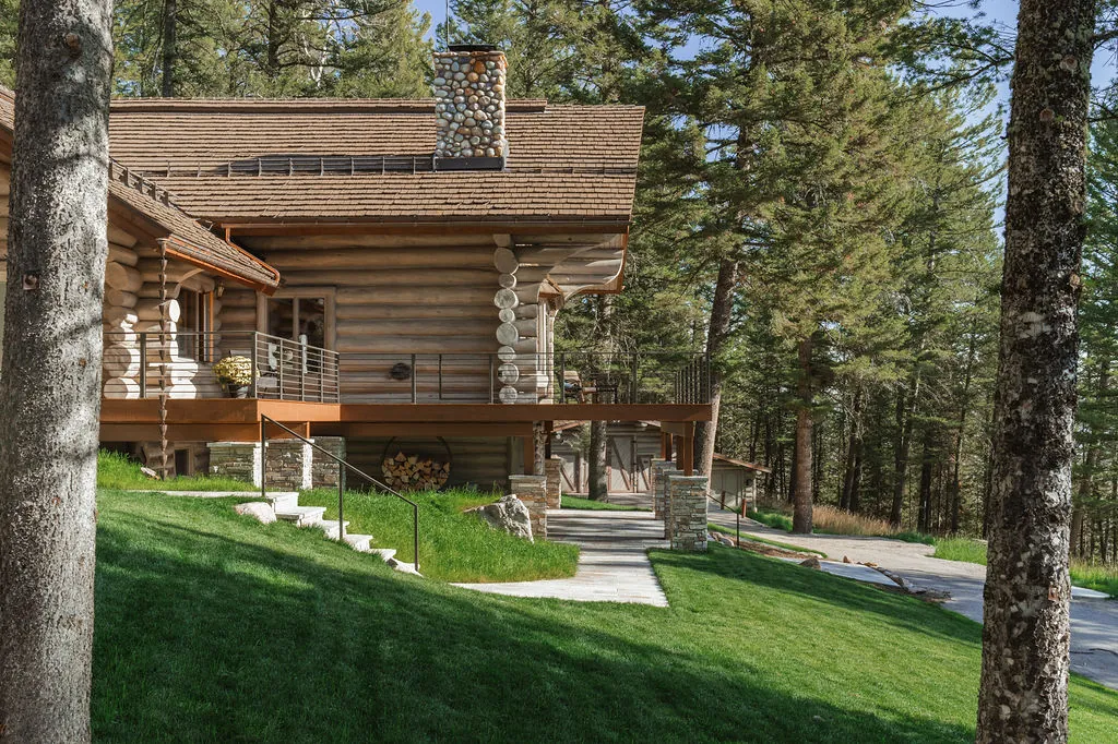A log home with a river-rock chimney and a long deck, on a green lawn among tall pines.